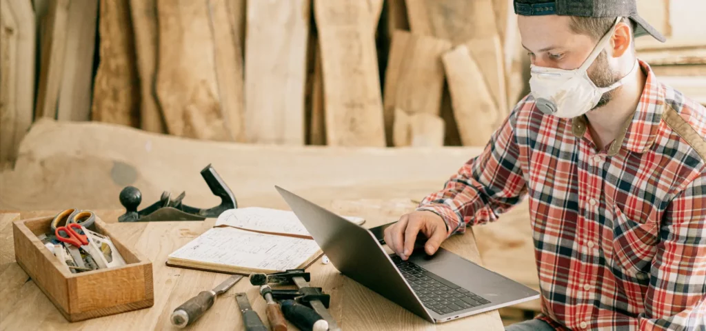 Ein Mann mit kariertem Hemd und Gesichtsmaske arbeitet an einem Laptop.